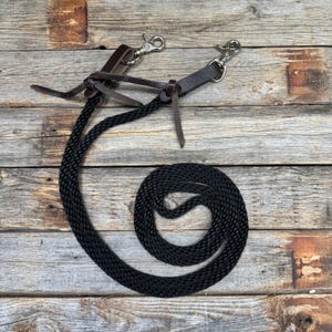 May include: Black nylon horse reins with leather detailing and silver-coloured metal clasps. The reins are coiled on a weathered wooden surface, showing the texture of the rope and leather.