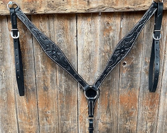 May include: Black leather horse breast collar with floral tooling and silver hardware. The collar is hanging on a wooden wall.