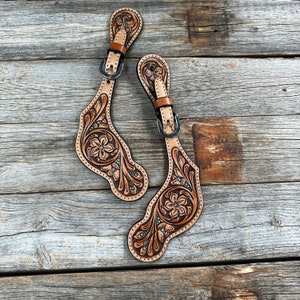 May include: A pair of brown leather spur straps with floral tooling and turquoise accents. The straps have silver buckles and are designed for cowboy boots.