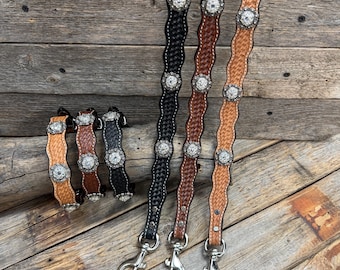 May include: Leather tack straps in various colors, including black, brown, and tan, with decorative silver-toned embellishments. The straps have a woven or textured design and are displayed on a wooden surface. Each strap has a metal clasp.