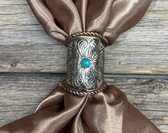 May include: A silver scarf ring with an embossed floral design and a turquoise center stone. The ring is wrapped around a brown satin scarf, creating a decorative accessory. The scarf and ring are displayed on a wooden surface.