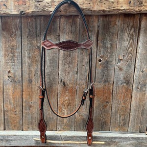 May include: A brown leather horse headstall with black stitching and silver hardware. The headstall is hanging on a wooden fence.