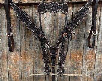 May include: A brown leather horse bridle with a decorative stitched pattern and copper accents. The bridle is hanging on a wooden wall.
