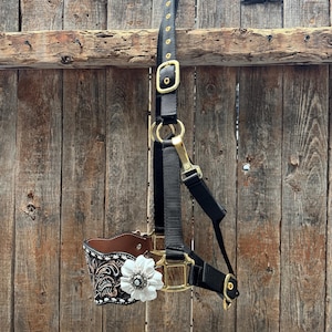May include: Black and gold horse halter with a brown leather noseband. The noseband features a black and white floral design and a white flower with a silver centre. The halter is hanging against a wooden fence.
