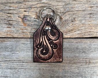 May include: A brown leather keychain with a silver key ring. The keychain features an embossed floral design and a silver rivet. The leather has a dark brown color with lighter brown accents. The keychain is rectangular with a rounded top.