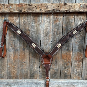 May include: A brown leather horse breast collar with white stitching and a diamond pattern. The collar has silver studs and is decorated with white arrow designs.