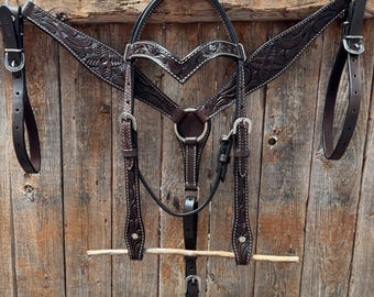 Puede incluir: Un conjunto de brida y petral de cuero marrón oscuro para caballos. El conjunto presenta intrincados grabados florales, costuras blancas y hebillas plateadas. La brida incluye una testera, carrilleras y una ahogadero. El petral tiene correas ajustables.