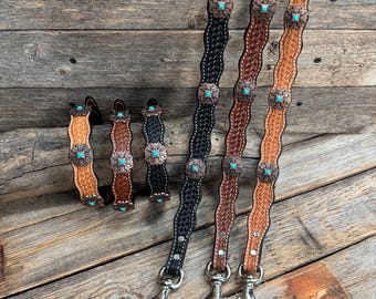 May include: Leather straps in various shades of brown and black, adorned with turquoise-colored accents and decorative metal flower-shaped embellishments. The straps have silver-colored metal clasps. The leather straps are displayed on a rustic wooden surface.