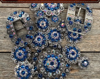 May include: A collection of silver-toned metal belt buckles and circular conchos embellished with vibrant blue and iridescent Swarovski crystals. The items feature intricate engraved patterns and the text CREATED IN USA WITH SWAROVSKI CRYSTALS.