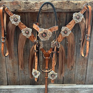 May include: A Western-style horse tack set, featuring a bridle, breast collar, and reins. The set is crafted from light brown leather, adorned with floral embellishments, red accents, and fringe. The background is a rustic wooden wall.