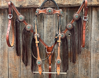 May include: A brown leather horse bridle with turquoise accents and fringe. The bridle is hanging on a wooden fence.