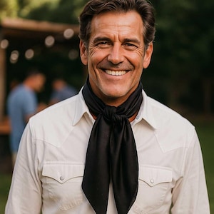 May include: A black neck scarf with a subtle dotted pattern is knotted at the neck. The scarf is paired with a light-colored button-down shirt with two chest pockets. The background is blurred, suggesting an outdoor setting.