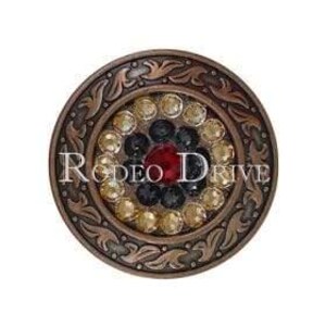 May include: A round, bronze-colored decorative button with a central red gem surrounded by black, gold, and clear gems. The button has an ornate, swirling border and the text "RODEO DRIVE" in white.