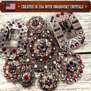 May include: A collection of metal belt buckles and circular conchos embellished with red, black, and champagne Swarovski crystals. The items feature intricate engraved patterns and geometric metalwork. Text at the top reads: CREATED IN USA WITH SWAROVSKI CRYSTALS.