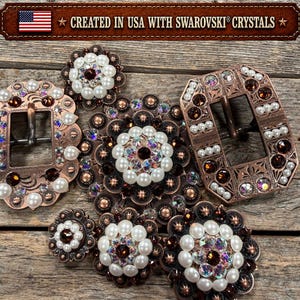 May include: A collection of copper-toned metal belt buckles and circular conchos embellished with white faux pearls and iridescent Swarovski crystals. The items feature intricate engraved patterns and are labelled as created in the USA.
