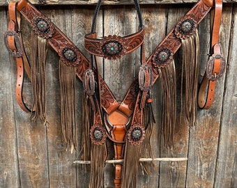 May include: A brown leather horse tack set with turquoise accents. The set includes a bridle, breast collar, and reins. The leather is tooled with floral designs and has fringe.