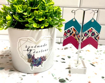 May include: Teal, pink, and cream layered leather earrings with a cheetah print pattern. The earrings are on a white stand with a green plant in a white pot. The pot has a sticker that says "handmade by darlene".