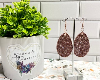 May include: A pair of brown leather earrings with a sparkly finish. The earrings are oval shaped and have a rose gold hook. The earrings are hanging from a clear stand. The text "handmade by darlene" is visible on a white pot with a green plant.