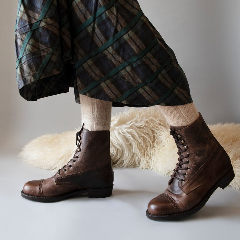 May include: Brown leather lace-up boots with a cap toe design. The boots are worn by a person wearing a plaid skirt and socks.