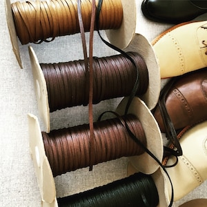Puede incluir: Cuatro carretes de cordón de cuero en colores marrón, beige y negro. Los cordones están enrollados alrededor de carretes de cartón con una etiqueta de papel blanco. Los carretes están dispuestos verticalmente en una fila.