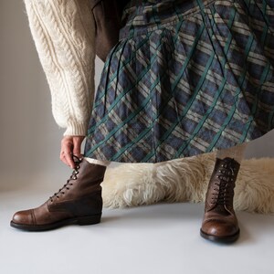 May include: A pair of brown leather lace-up boots with a chunky heel. The boots are worn with a plaid skirt and a white sweater.