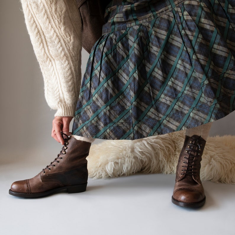 May include: A pair of brown leather lace-up boots with a chunky heel. The boots are worn with a plaid skirt and a white sweater.