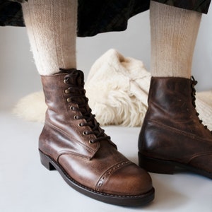 May include: A pair of brown leather lace-up boots with a cap toe design. The boots have a thick sole and are worn with white socks.