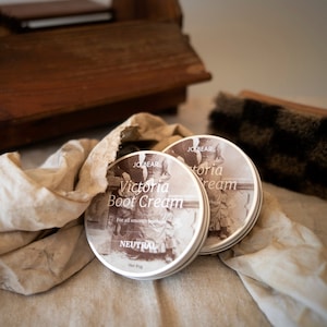 May include: Two round tins of "Victoria Boot Cream" with a vintage label, resting on a beige cloth. The tins are labeled "Neutral" and "For all smooth leathers". A wooden shoe shine box and brush are in the background.