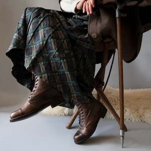 May include: A pair of brown leather boots with laces. The boots have a pointed toe and a chunky heel. The boots are worn by a person sitting on a wooden chair with a plaid skirt.
