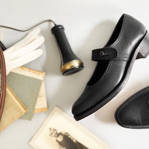 Op de afbeelding: Een paar zwarte leren schoenen met een band en gesp. De schoenen hebben een lage hak en staan op een wit oppervlak.