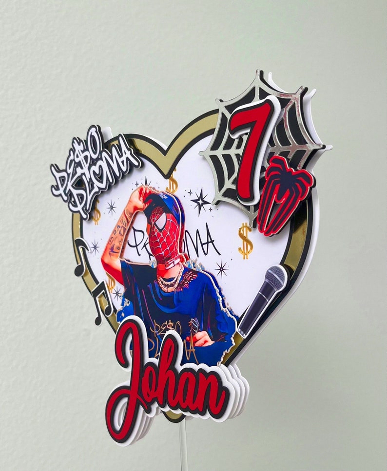 Puede incluir: Un topper de pastel de Spiderman rojo y negro con un fondo en forma de coraz&oacute;n. El topper presenta un personaje de Spiderman, un micr&oacute;fono, el n&uacute;mero 7 y el nombre "Johan".
