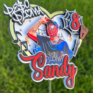 Puede incluir: Un topper de pastel rojo y blanco con un personaje de dibujos animados que lleva una m&aacute;scara de Spiderman roja y azul, una camisa azul con las palabras "Peso Pluma" en blanco y una cadena de oro. El topper tambi&eacute;n incluye un coraz&oacute;n blanco con detalles dorados, una nota musical y la palabra "Sandy" en rojo y negro.