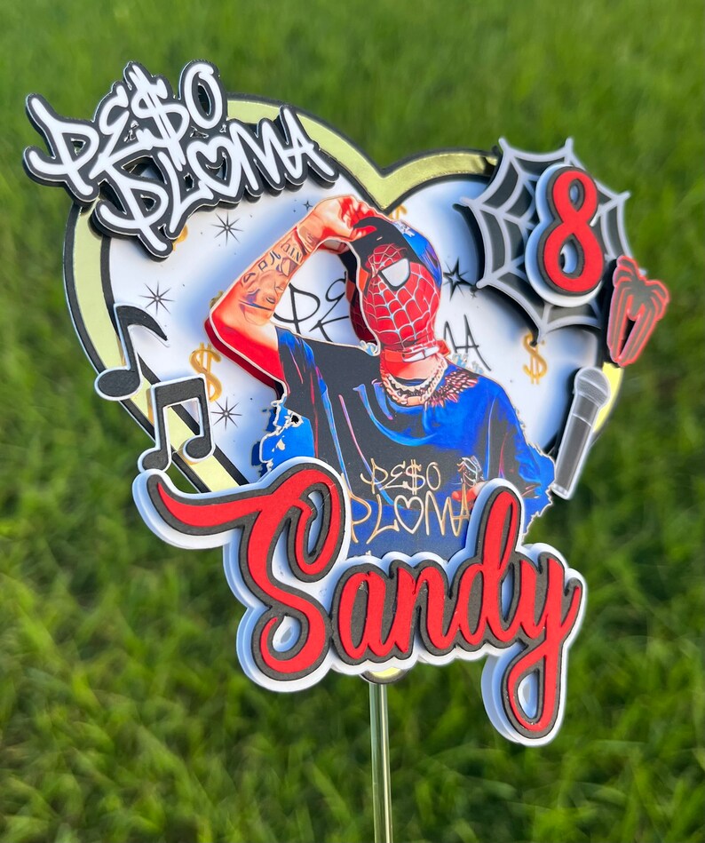 Puede incluir: Un topper de pastel rojo y blanco con un personaje de dibujos animados que lleva una m&aacute;scara de Spiderman roja y azul, una camisa azul con las palabras "Peso Pluma" en blanco y una cadena de oro. El topper tambi&eacute;n incluye un coraz&oacute;n blanco con detalles dorados, una nota musical y la palabra "Sandy" en rojo y negro.