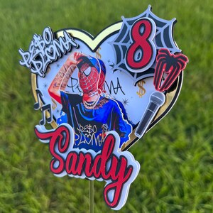 Puede incluir: Un adorno para pastel rojo y negro con una base en forma de coraz&oacute;n, que presenta un personaje de dibujos animados con un disfraz de Spiderman rojo y azul, un micr&oacute;fono, una telara&ntilde;a con el n&uacute;mero 8 y el texto "Peso Pluma" y "Sandy".