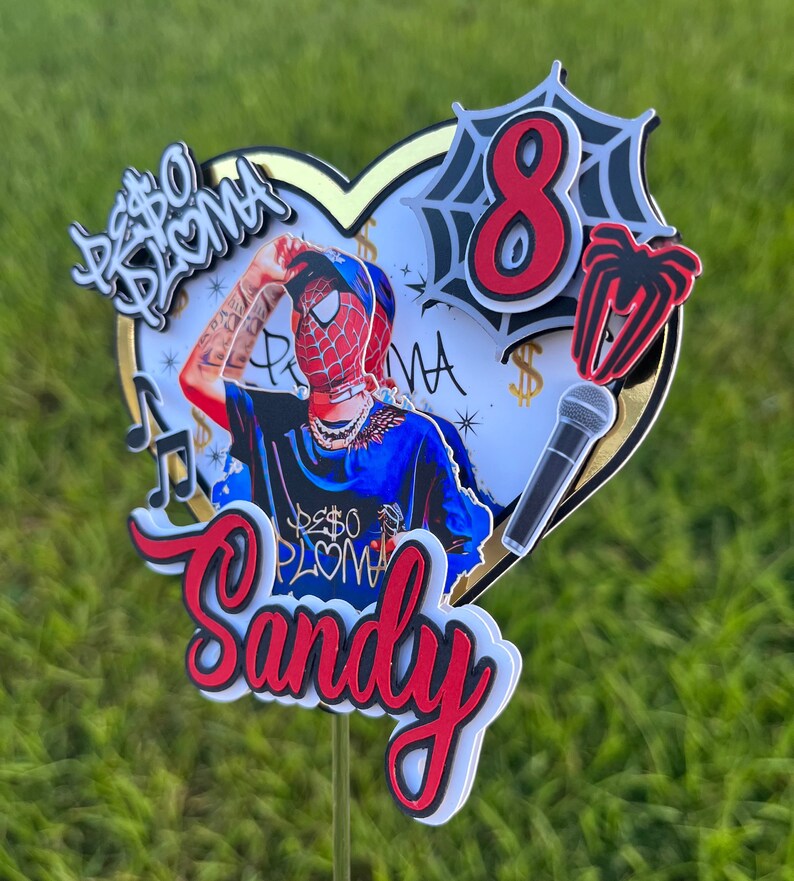 Puede incluir: Un adorno para pastel rojo y negro con una base en forma de coraz&oacute;n, que presenta un personaje de dibujos animados con un disfraz de Spiderman rojo y azul, un micr&oacute;fono, una telara&ntilde;a con el n&uacute;mero 8 y el texto "Peso Pluma" y "Sandy".