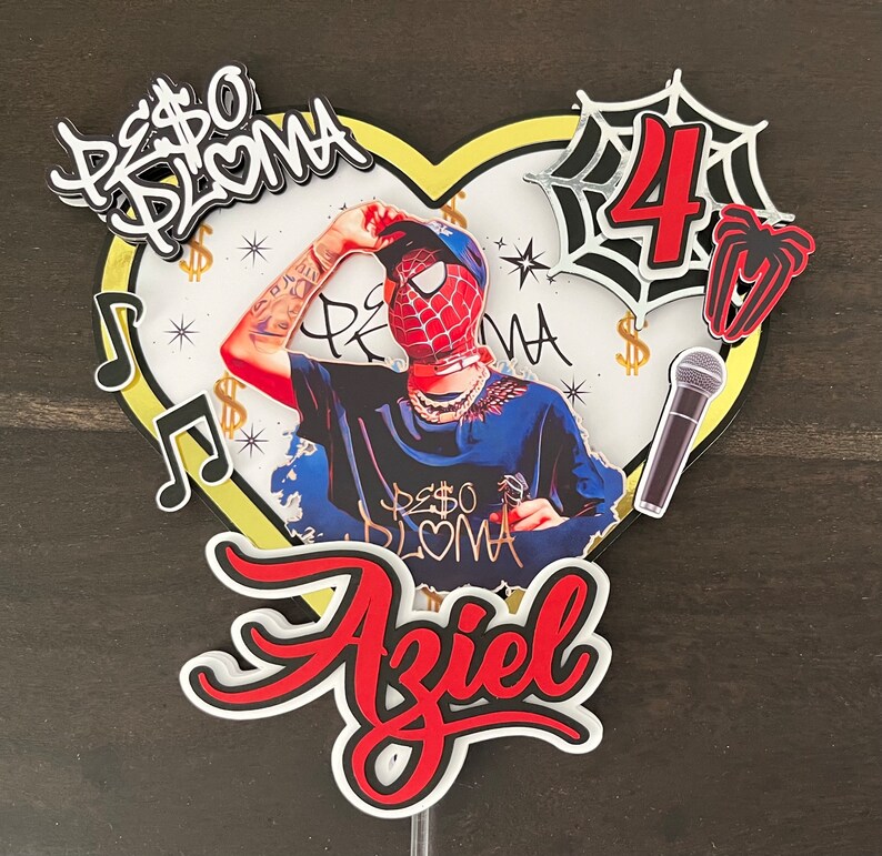 Puede incluir: Un adorno para pastel rojo, blanco y negro con un personaje de dibujos animados que lleva una m&aacute;scara de Spider-Man roja y azul, un micr&oacute;fono blanco y negro, una telara&ntilde;a blanca y negra con el n&uacute;mero 4 en el centro y el nombre Aziel en letras rojas y negras.