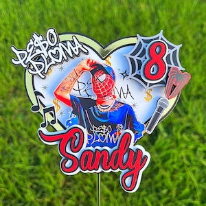 Puede incluir: Un topper de pastel rojo, blanco y negro con una base en forma de coraz&oacute;n. El topper presenta una imagen de dibujos animados de una persona con una m&aacute;scara de Spider-Man, una cadena de oro y una camisa azul. El texto "Peso Pluma" est&aacute; escrito en blanco y negro en el topper. El n&uacute;mero "8" est&aacute; escrito en rojo y blanco con una telara&ntilde;a negra detr&aacute;s. El nombre "Sandy" est&aacute; escrito en rojo y blanco con un contorno negro.