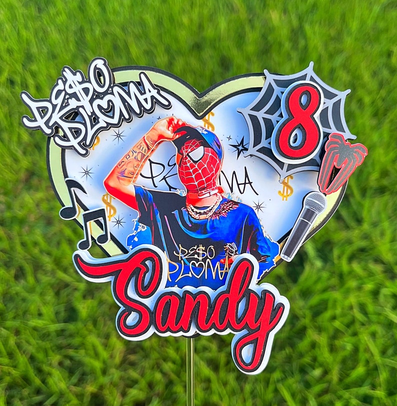 Puede incluir: Un topper de pastel rojo, blanco y negro con una base en forma de coraz&oacute;n. El topper presenta una imagen de dibujos animados de una persona con una m&aacute;scara de Spider-Man, una cadena de oro y una camisa azul. El texto "Peso Pluma" est&aacute; escrito en blanco y negro en el topper. El n&uacute;mero "8" est&aacute; escrito en rojo y blanco con una telara&ntilde;a negra detr&aacute;s. El nombre "Sandy" est&aacute; escrito en rojo y blanco con un contorno negro.