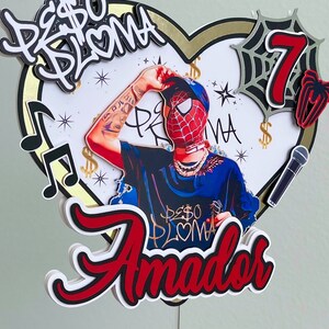 Puede incluir: Un topper de pastel rojo, blanco y negro con un personaje de dibujos animados que lleva una m&aacute;scara roja y azul, una cadena de oro y una camisa negra. El topper tambi&eacute;n incluye un micr&oacute;fono, una telara&ntilde;a y el texto "Peso Pluma" y "Amador".