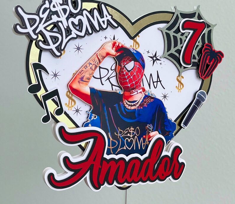 Puede incluir: Un topper de pastel rojo, blanco y negro con un personaje de dibujos animados que lleva una m&aacute;scara roja y azul, una cadena de oro y una camisa negra. El topper tambi&eacute;n incluye un micr&oacute;fono, una telara&ntilde;a y el texto "Peso Pluma" y "Amador".