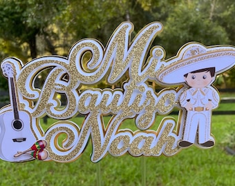 May include: Gold glitter cake topper with the words "Mi Bautizo Noah" and a cartoon image of a boy wearing a sombrero and a white suit holding a guitar and maracas.