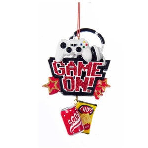 May include: A red and black Christmas ornament with the words "Game On!" and a video game controller, headphones, a can of soda, and a bag of chips.