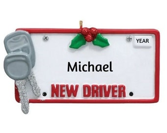 Può includere: Un ornamento natalizio rosso e bianco a forma di targa con le parole "NEW DRIVER" e un ramo di agrifoglio. L'ornamento ha un ciondolo a forma di chiave argentata e uno spazio vuoto per un nome, che è riempito con il nome "Michael".