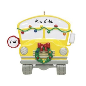 Può includere: Addobbo natalizio a forma di autobus scolastico giallo con un fiocco rosso e una ghirlanda verde. L'autobus ha un cartello bianco sul davanti che dice "Mrs. Kidd" e un cartello di stop rosso che dice "Anno".