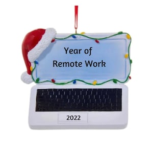 Op de afbeelding: Witte laptop kerstversiering met een rode kerstmanmuts op de bovenkant. Het scherm zegt "Jaar van Remote Work" met kleurrijke lampjes rond de rand. Het jaar 2022 is onderaan gedrukt.