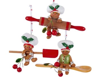 Pode incluir: Conjunto de três enfeites de gingerbread, cada um vestido como um padeiro e segurando uma ferramenta de cozinha diferente: um rolo, uma espátula e uma colher de pau. Cada enfeite é decorado com detalhes vermelhos e verdes e tem um chapéu de chef branco com azevinho.
