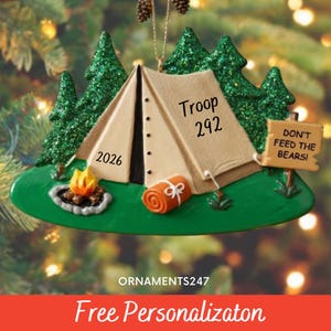 May include: A camping-themed Christmas ornament featuring a tan tent with the text Troop 292 and 2026, a small campfire, a rolled orange sleeping bag, and a sign reading DON'T FEED THE BEARS, all set against a background of green glittery pine trees.