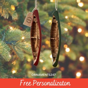 May include: Two canoe-shaped Christmas ornaments. One is red, the other green, both with a wooden interior and a small paddle. A tag that says "Handmade" hangs from the red ornament. The background is a blurred Christmas tree with lights.