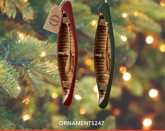 Könnte beinhalten: Zwei weihnachtliche Kanu-Ornamente, eines rot und eines grün, beide mit Holzinnenraum und kleinem Paddel. Ein Anhänger mit der Aufschrift "Handmade" hängt am roten Ornament. Der Hintergrund ist ein verschwommener Weihnachtsbaum mit Lichtern.