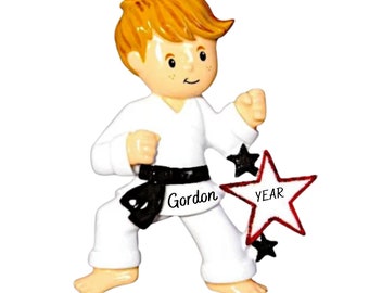 May include: A white karate uniform ornament with a black belt, a red star with the word "YEAR" on it, and the name "Gordon" on the belt. The ornament is of a boy with blond hair.